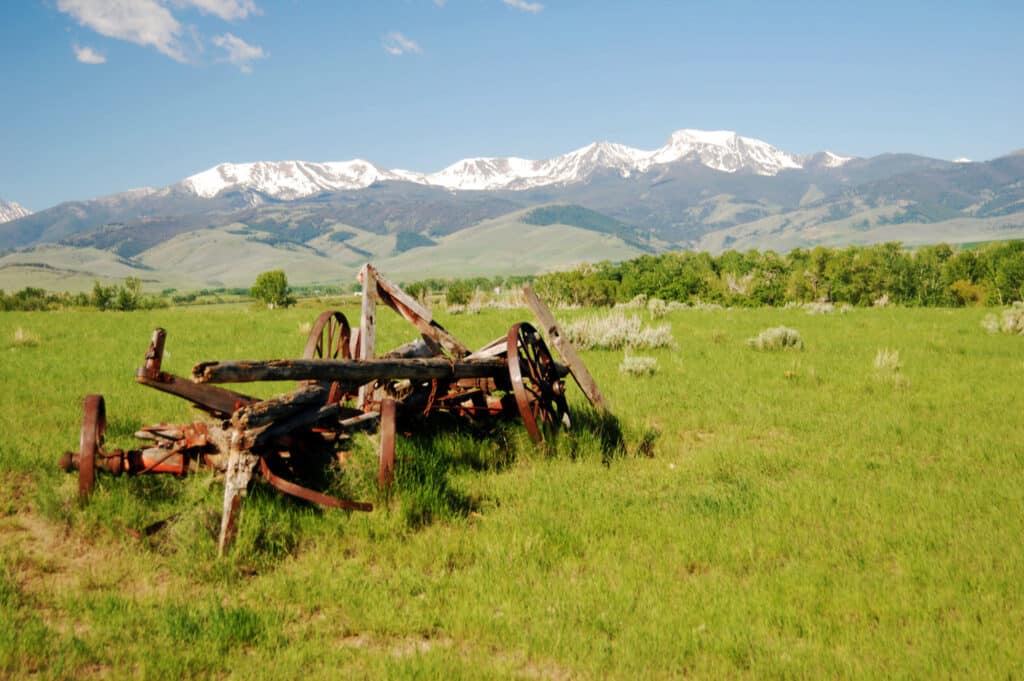 Rusty, broken-down farm equipment sits in a green grassy field on a cattle ranch with distant mountains, partially covered in snow, under a clear blue sky.