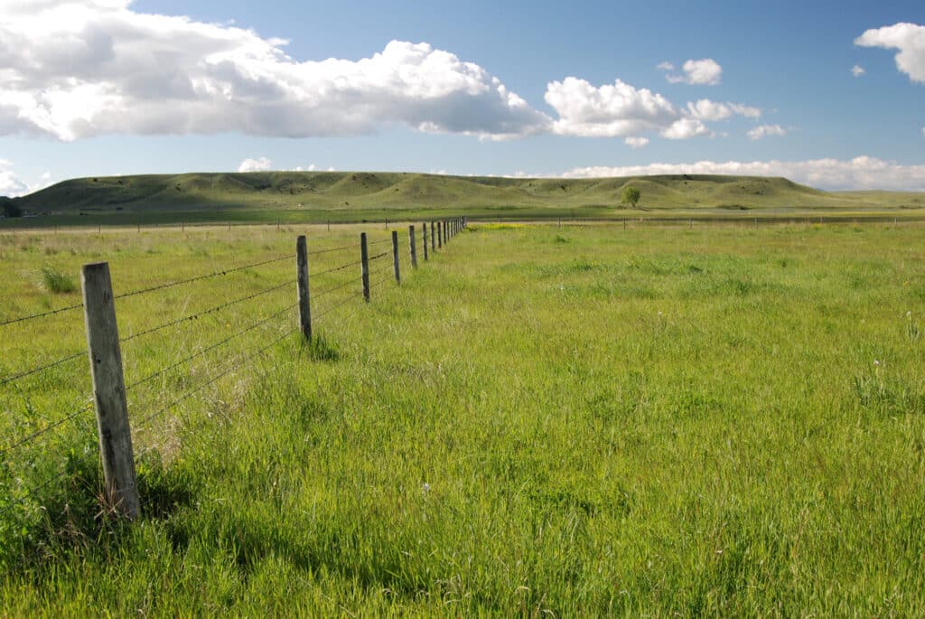 A wire fence stretches across a green grassy field under a blue sky with scattered clouds and low rolling hills—ideal recreational land or cattle ranch, with scenic views in every direction.