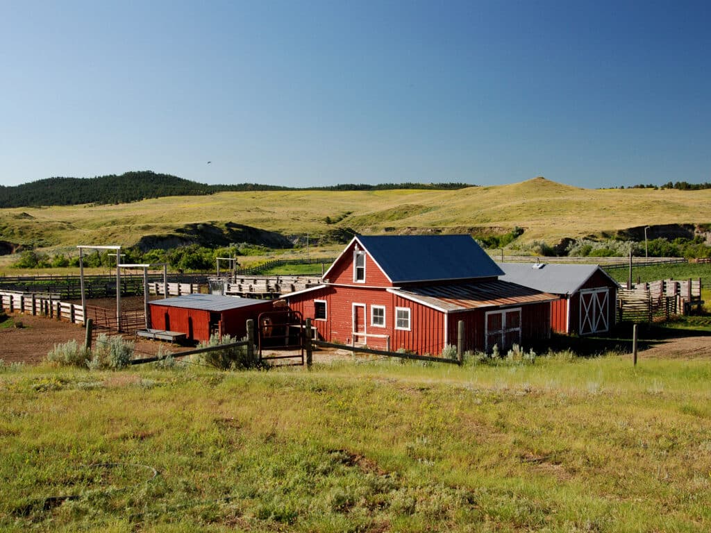 A red barn with white trim sits in the middle of a fenced area on a grassy farm, perfect as recreational land or hunting property, with rolling green hills and a clear blue sky in the background.
