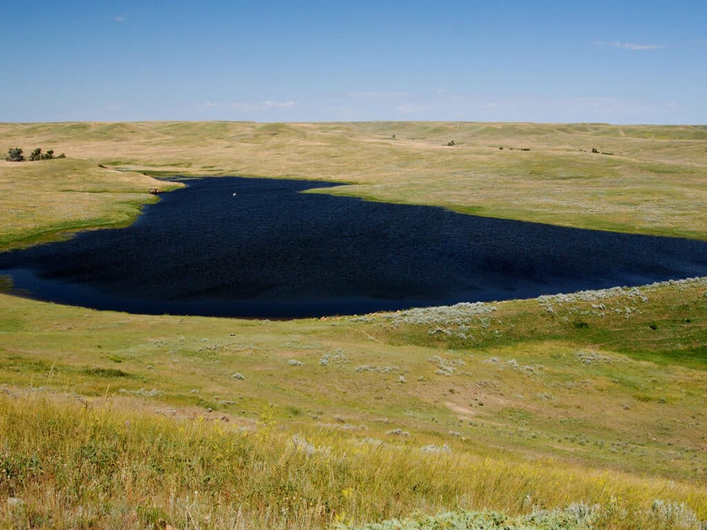 A small, dark blue lake surrounded by open, rolling grasslands on recreational land under a clear blue sky, with sparse vegetation and gentle hills in the background—perfect for a serene cattle ranch setting.