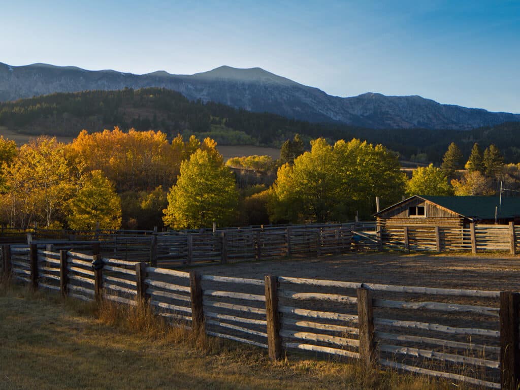 A wooden fence encloses a pasture with a rustic barn, surrounded by trees in autumn colors. In the background, mountains rise under a clear blue sky—ideal land for sale as a scenic cattle ranch or hunting property.
