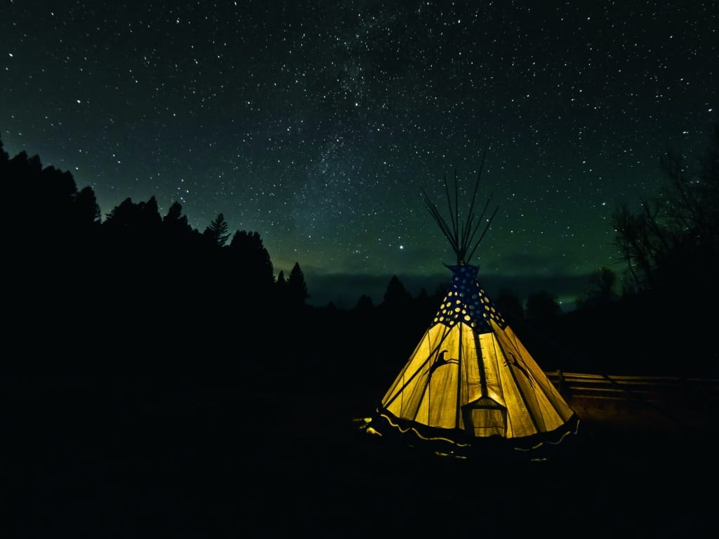 A warmly lit tipi stands in the foreground under a clear, star-filled night sky on recreational land, surrounded by silhouetted trees and a dark landscape.