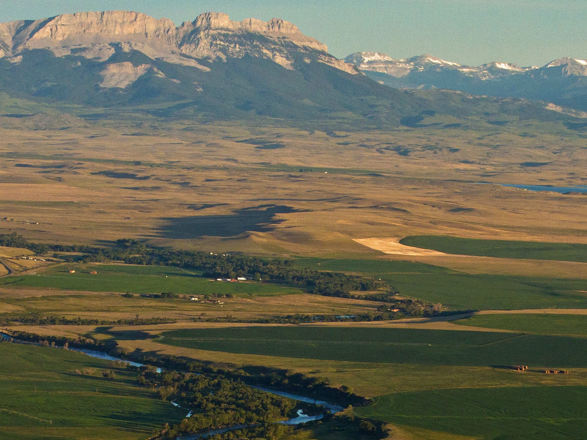 Wide view of a green and golden prairie landscape with a winding river, fields, scattered farm buildings, and cattle ranch. Rugged mountains with rocky peaks and some snow rise in the distance under a clear sky.