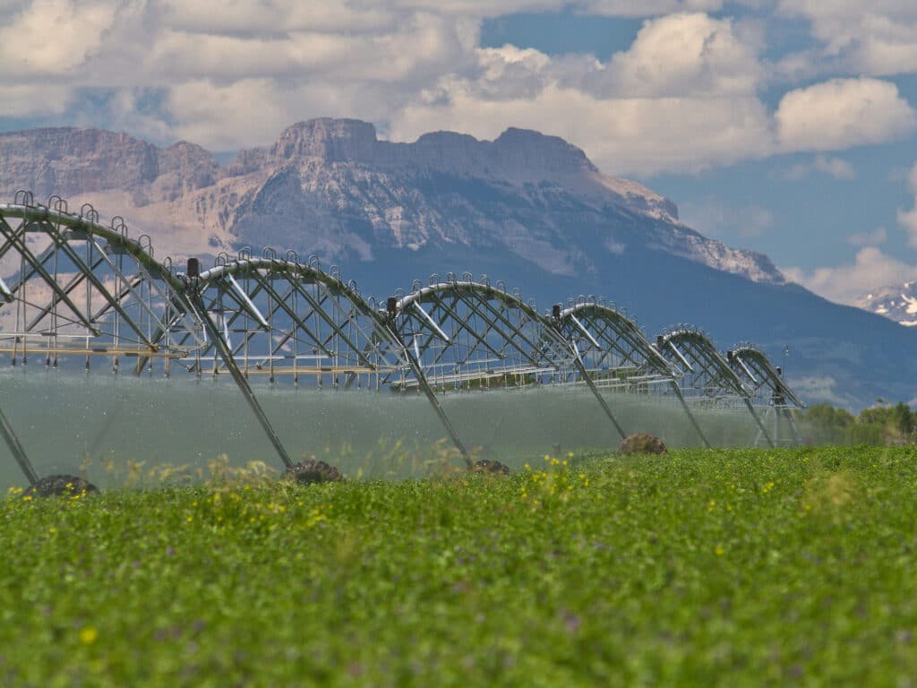 A large irrigation system sprays water over a green agricultural field on recreational land, with mountains and a partly cloudy sky in the background.
