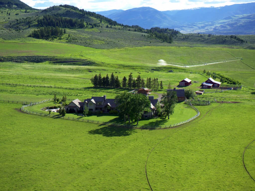 Aerial view of a cattle ranch with several buildings surrounded by green fields, trees, and distant hills under a blue sky with scattered clouds. A wooden fence encloses this ideal recreational land.