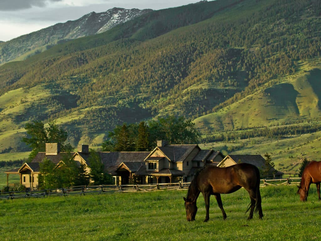 A brown horse grazes on green grass in front of a rustic house on a cattle ranch, with a wooden fence and tree-lined mountains in the background under a cloudy sky.