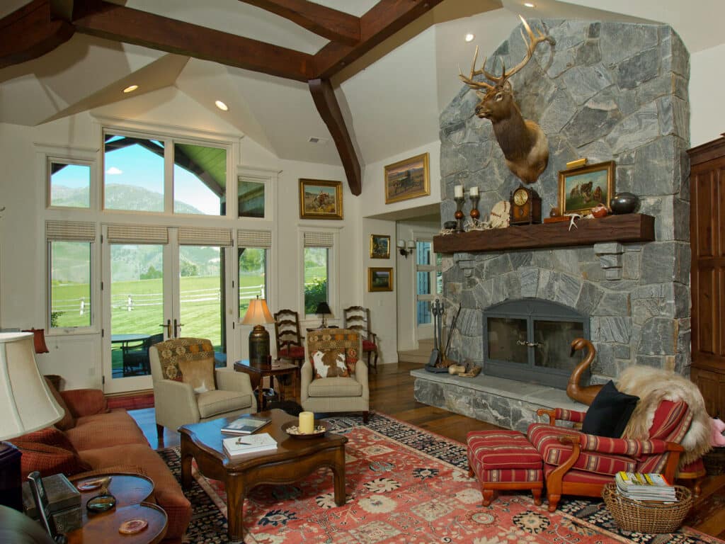 A cozy living room with a high-beamed ceiling, stone fireplace with a mounted deer head, patterned rug, wooden furniture, art on the walls, and large windows overlooking a scenic green landscape on recreational land.