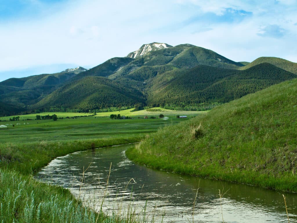 A winding stream flows through green grass on scenic recreational land, with tree-covered mountains and patches of snow in the background under a partly cloudy sky.