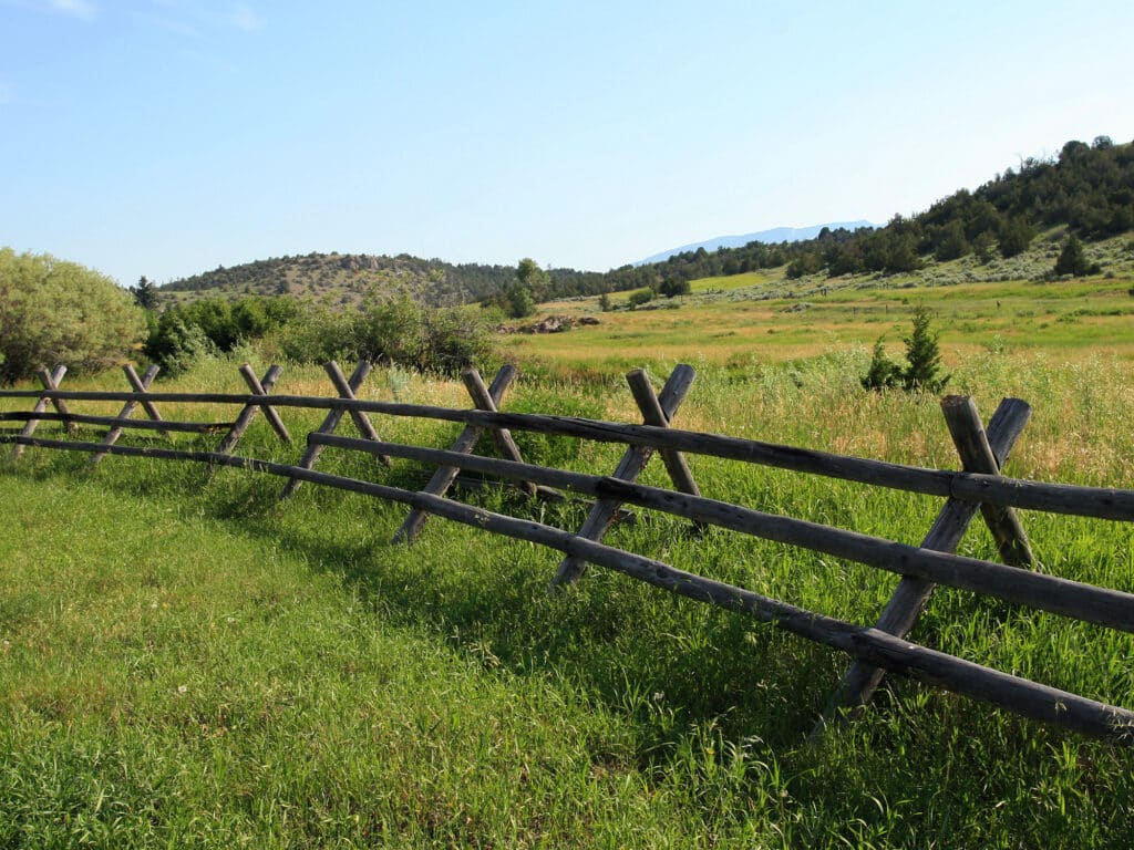 A rustic wooden fence runs diagonally through a grassy field, perfect for recreational land or a hunting property, with green hills and trees in the background under a clear blue sky.