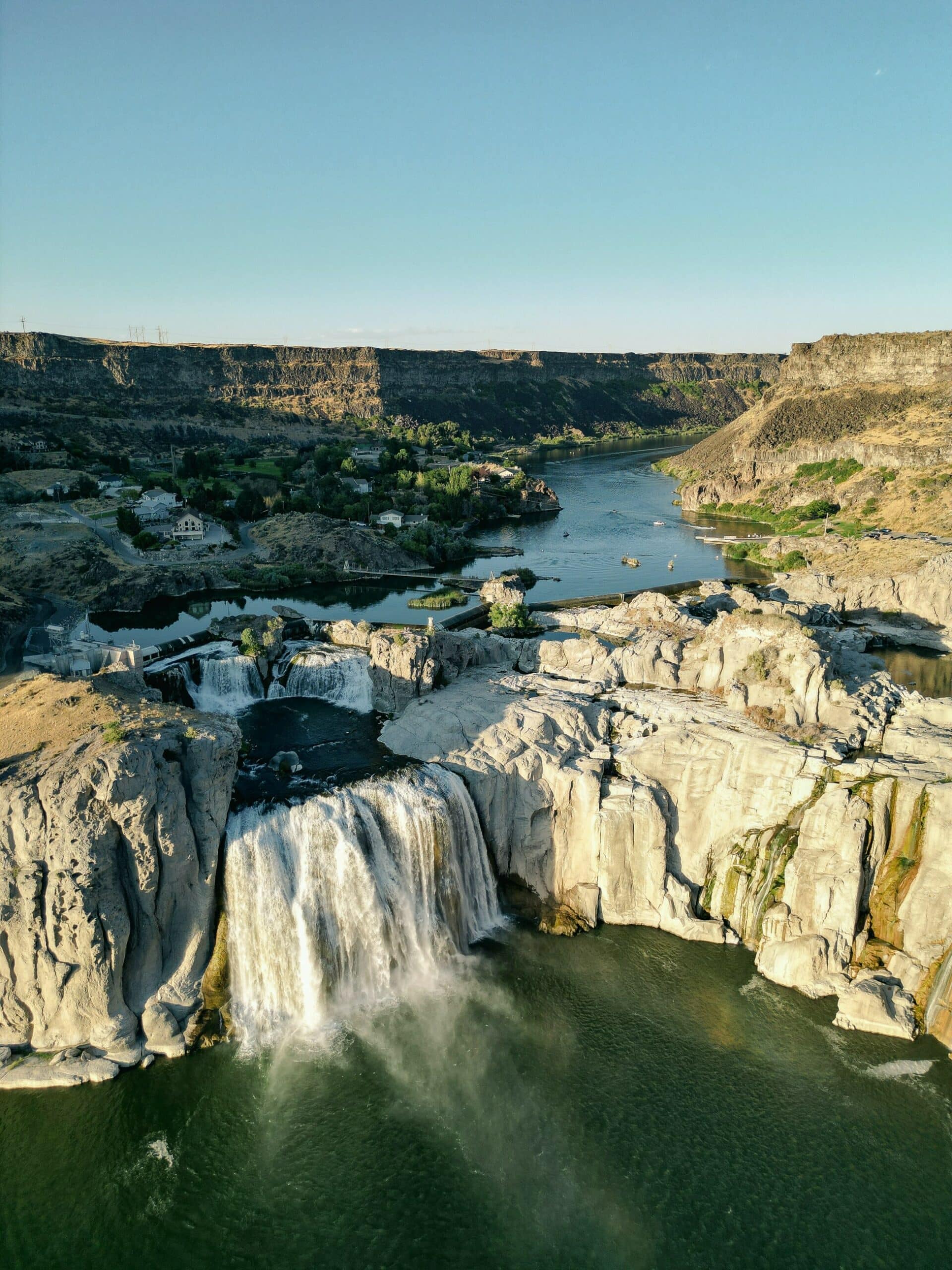 A wide waterfall cascades over rocky cliffs into a river, with green vegetation, scattered buildings, and rugged canyon walls under a clear sky—an idyllic setting perfect for a cattle ranch or prime land for sale.