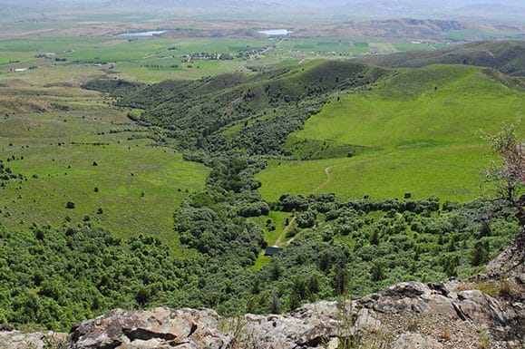 View from a rocky ledge overlooking a lush green valley, rolling hills, dense patches of trees, distant farmland, and a small settlement—ideal recreational land or potential ranch for sale under a hazy sky.