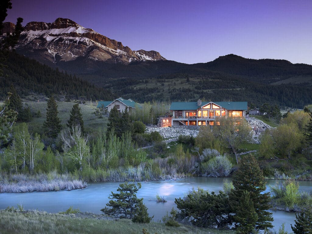 A large lodge with glowing windows sits in a lush valley beside a river on prime recreational land, surrounded by green trees and hills, with snow-capped mountains in the background under a twilight sky.