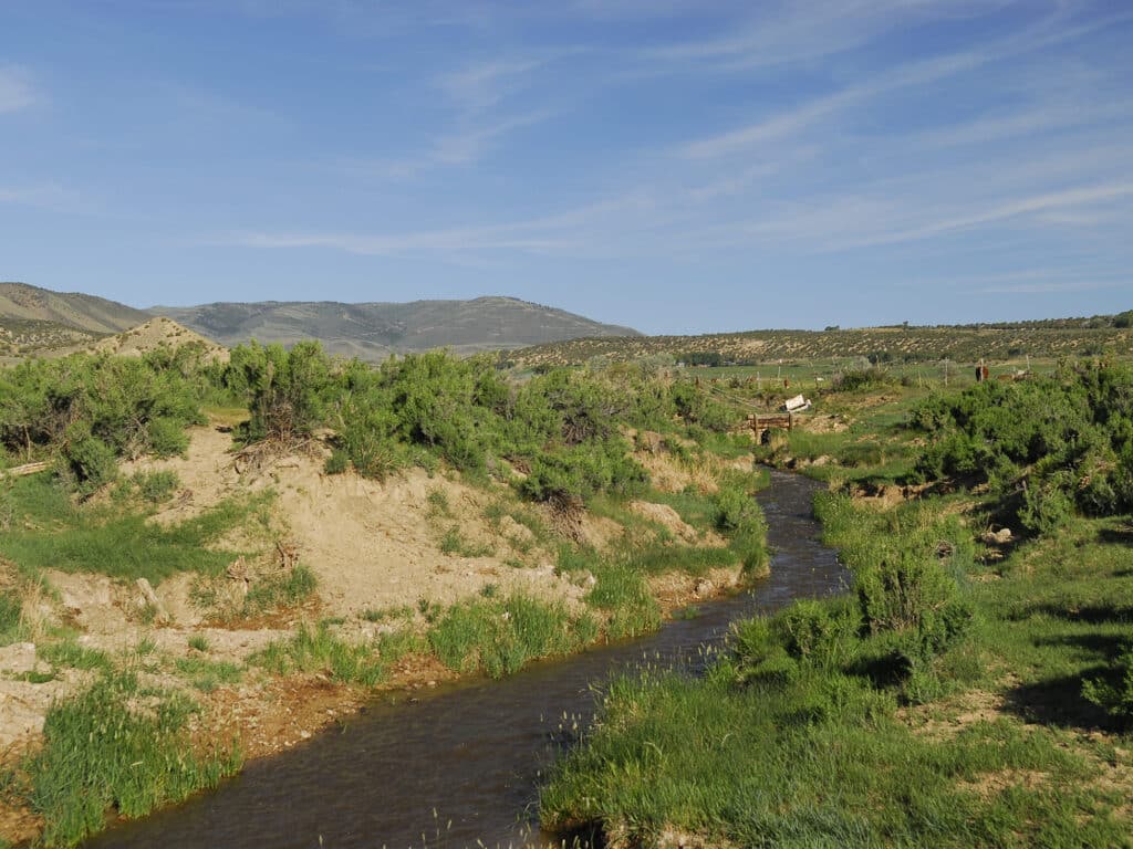 A narrow stream winds through green shrubs and grass under a blue sky with wispy clouds, perfect recreational land for sale with low hills and sparse vegetation in the background.