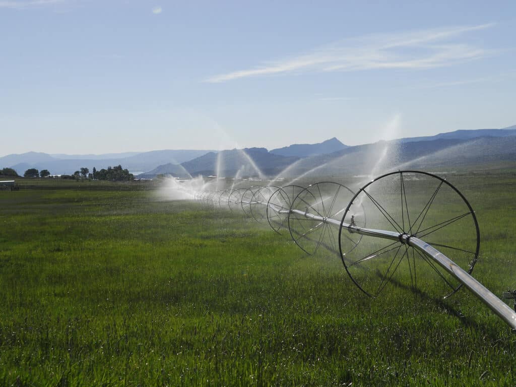 A large field on this ranch for sale is irrigated by a rotating sprinkler system with metal wheels, spraying water over green grass under a clear blue sky with distant mountains in the background.