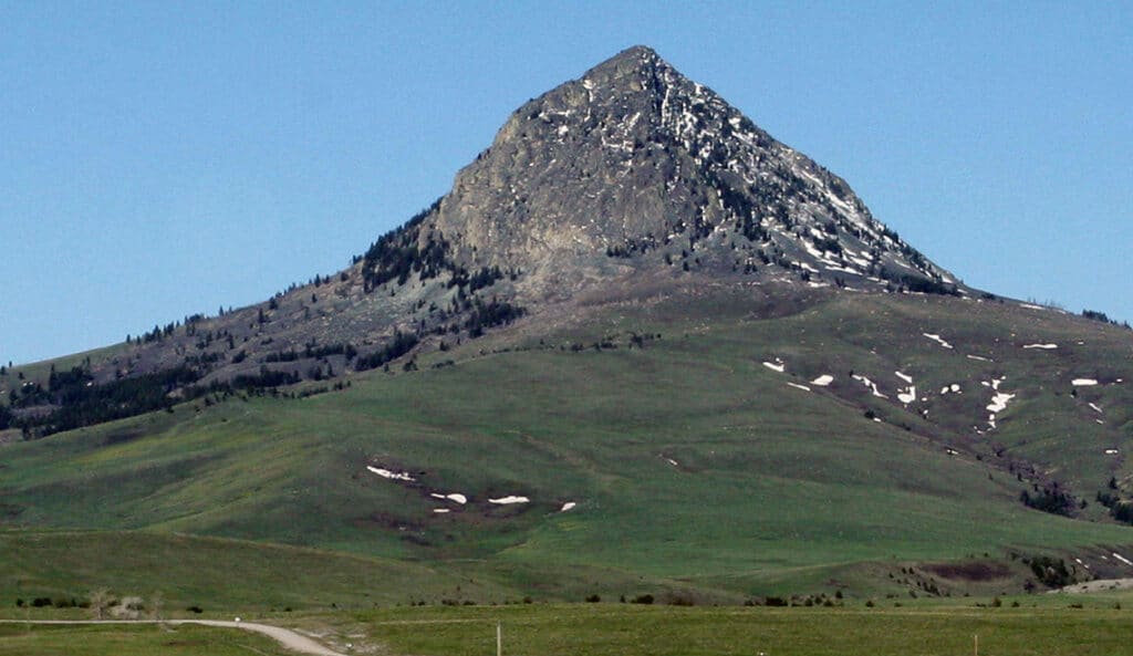 A prominent, rocky mountain peak rises above green, gently sloping hills—perfect land for sale—with patches of snow and sparse trees under a clear blue sky.
