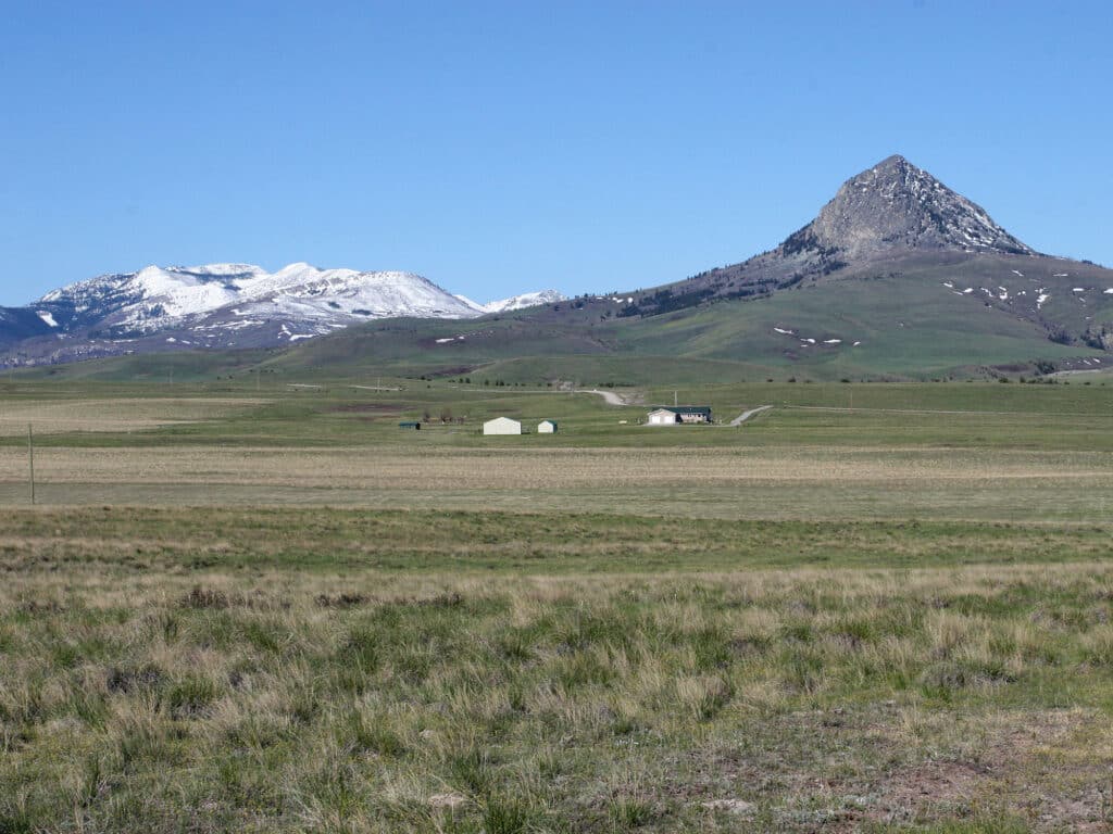 A grassy plain with scattered buildings sits beneath a pyramid-shaped mountain; snowy peaks rise in the background under a clear blue sky, offering an ideal cattle ranch or land for sale.