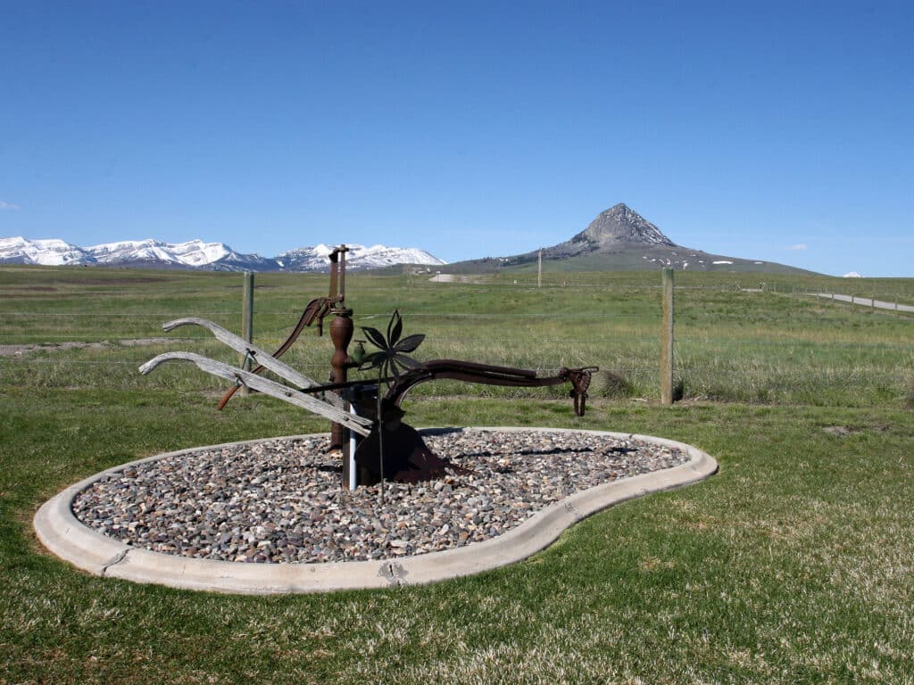 A metal sculpture resembling farm tools stands on a gravel bed in a grassy field, with snow-capped mountains and a distinct pointed peak in the background—an inspiring view for anyone seeking land for sale or envisioning their own cattle ranch.