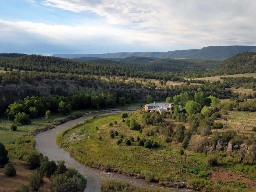 A winding river flows through a green valley with scattered trees and brush; a building complex sits near the water, surrounded by vegetation and hills under a cloudy sky—ideal as recreational land or a hunting property.