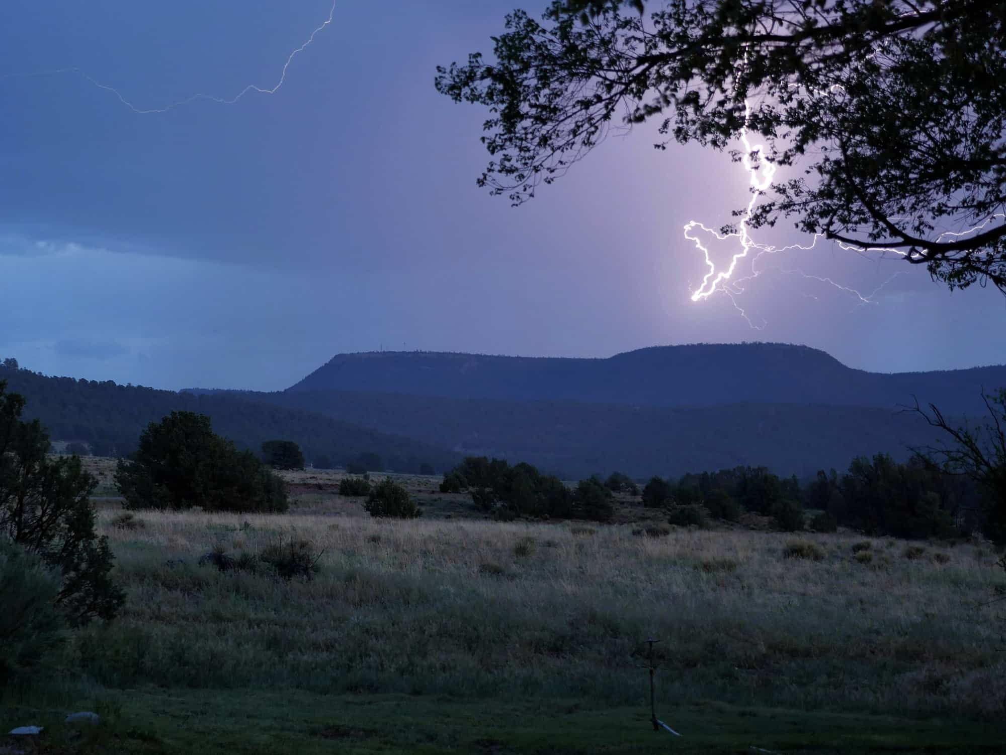A bolt of lightning strikes near a distant mesa at dusk, illuminating the cloudy sky over a grassy field with scattered trees—an impressive scene on this potential cattle ranch or hunting property.