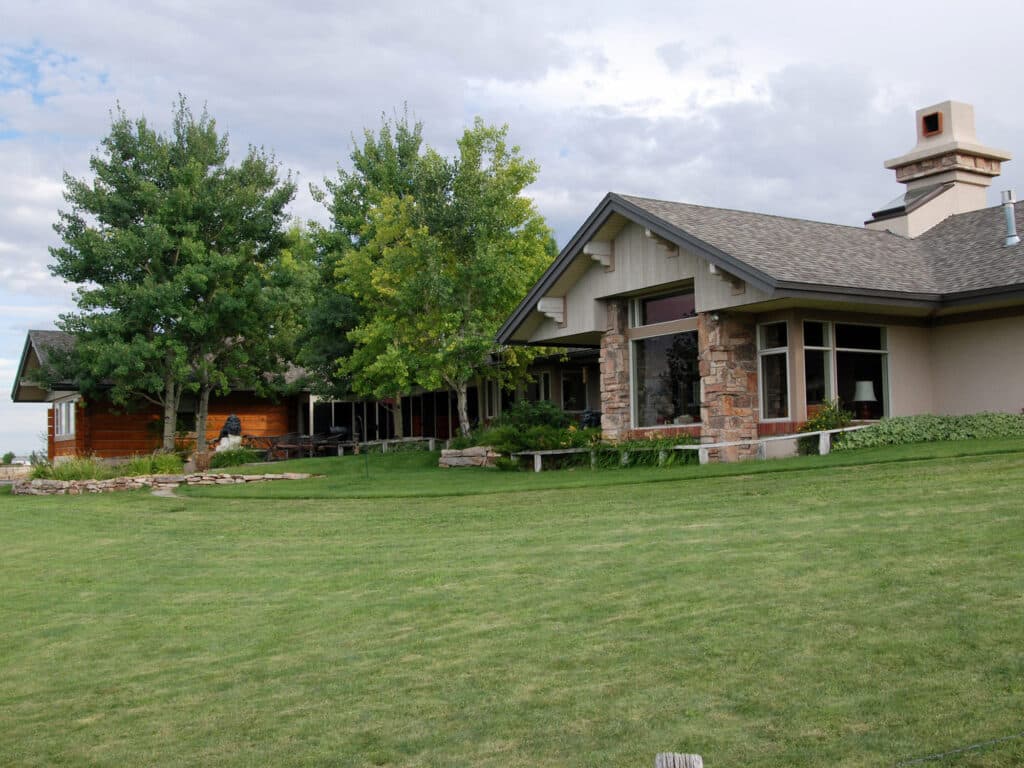 A large house with stone and wood exterior sits on a well-kept green lawn, surrounded by leafy trees under a partly cloudy sky—perfect as a ranch for sale or hunting property.