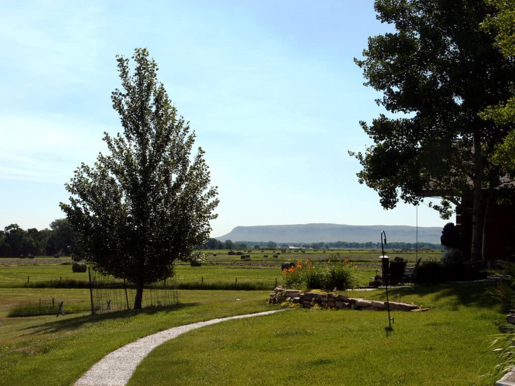 A curving path leads past a tree through a green lawn toward open fields, with hay bales and mountains visible in the distance under a clear blue sky—ideal hunting property or cattle ranch land for sale. A shaded porch is partially visible on the right.