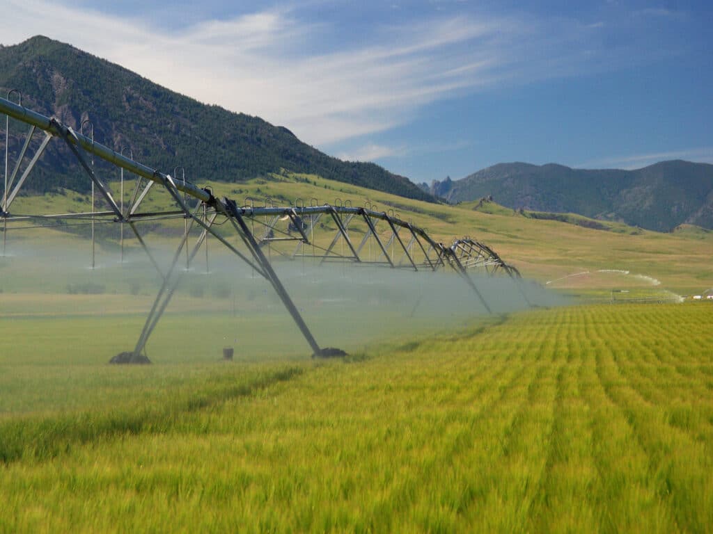 A large center pivot irrigation system sprays water over a lush green crop field, with mountains and a blue sky dotted with wispy clouds in the background—perfect for recreational land or a ranch for sale.