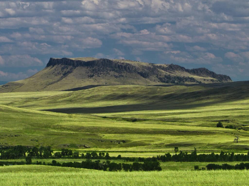 A broad, grassy plain stretches toward a flat-topped, rocky butte under a cloudy sky. Sunlight highlights patches of green fields, making this recreational land ideal for nature lovers or as a prime hunting property. Scattered shrubs and trees dot the landscape.
