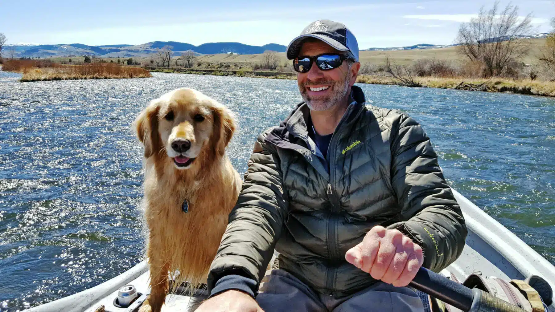 A man wearing sunglasses and a cap rows a boat on a river, smiling, with a golden retriever beside him. Mountains, grassy fields, and hints of hunting property are visible in the background under a clear sky.