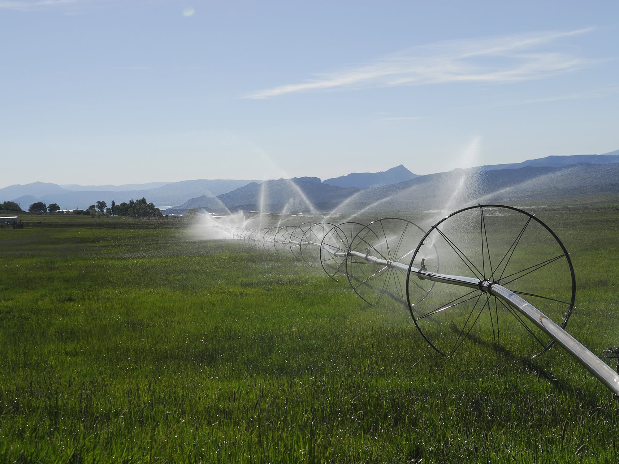 A long irrigation system with large wheels sprays water over a green grassy field under a clear sky, with distant mountains in the background—ideal recreational land or ranch for sale.