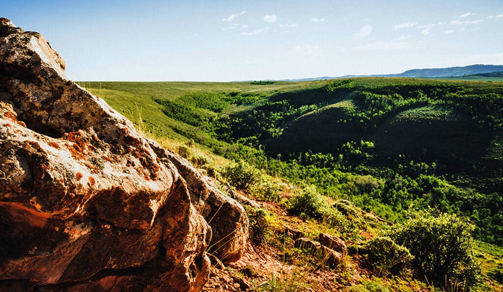 A large, sunlit rock sits in the foreground overlooking a lush, green valley—ideal cattle ranch or hunting property potential—with rolling hills covered in trees under a clear blue sky.