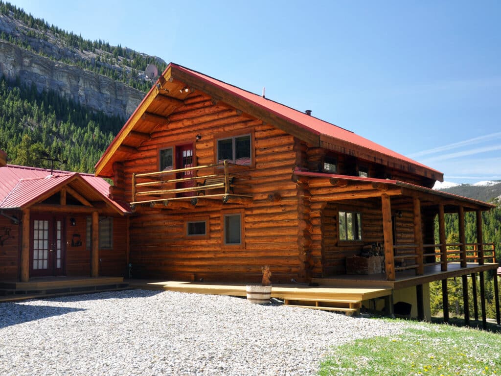 A large wooden log cabin with a red roof and wraparound porch sits on a gravel driveway, surrounded by pine trees and mountains under a clear blue sky—perfect as recreational land or your dream hunting property.