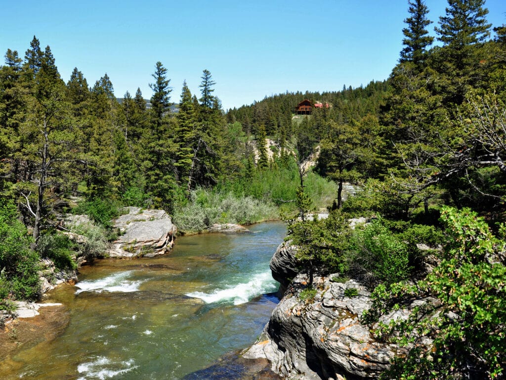 A clear river winds through rocky, forested recreational land with tall evergreens under a bright blue sky. A wooden cabin sits on a distant hill among the trees, perfect for those seeking unique hunting property.