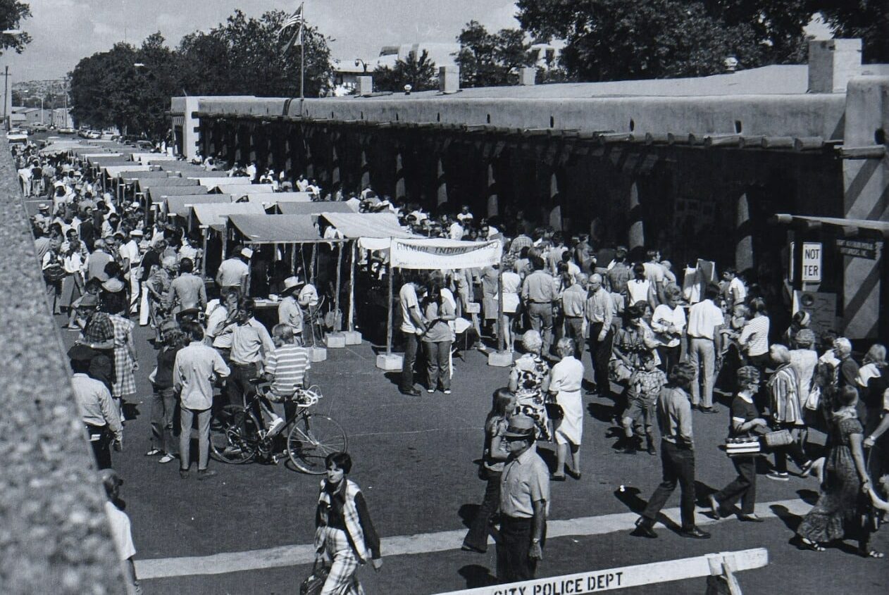 Black and white photo of a busy outdoor market with rows of stalls, large crowds browsing and walking, some people on bicycles, and a “City Police Dept” barricade in the foreground. Trees, buildings, and hints of recreational land line the background.