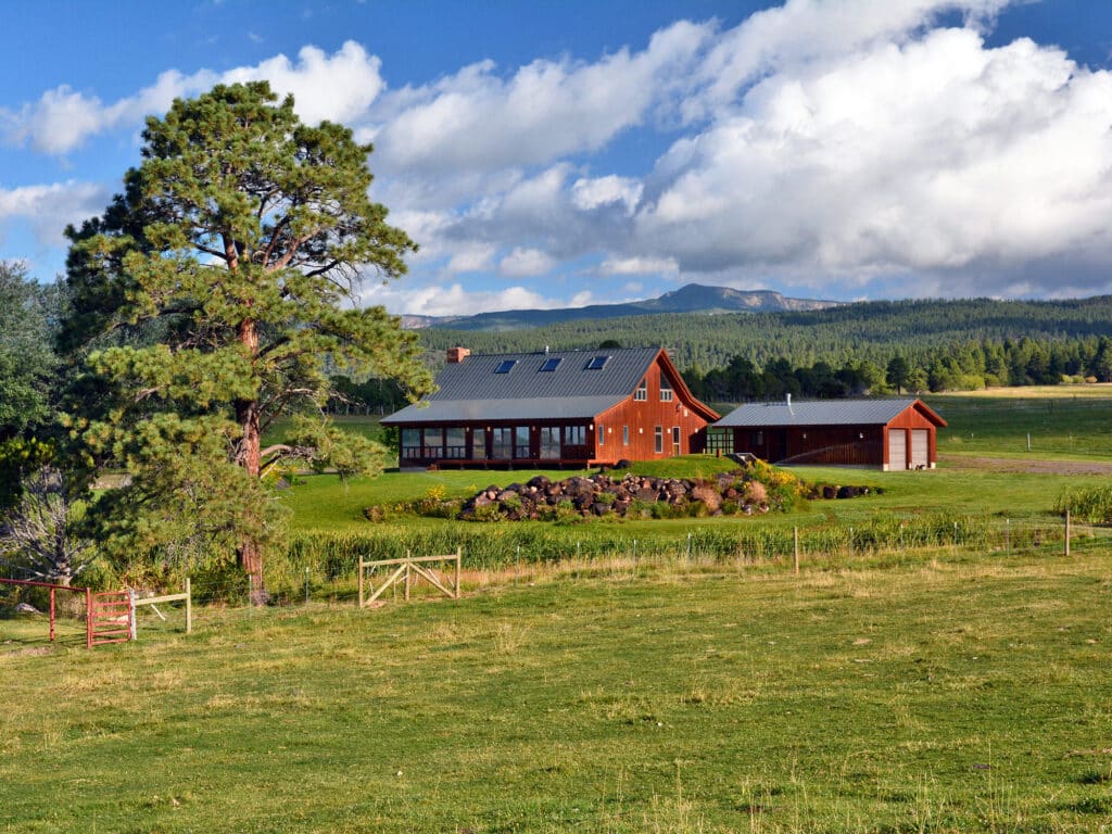 A red farmhouse with a gray roof sits in a green field, surrounded by trees and shrubs, with mountains and a partly cloudy blue sky in the background. This scenic hunting property features a wooden gate in the foreground.