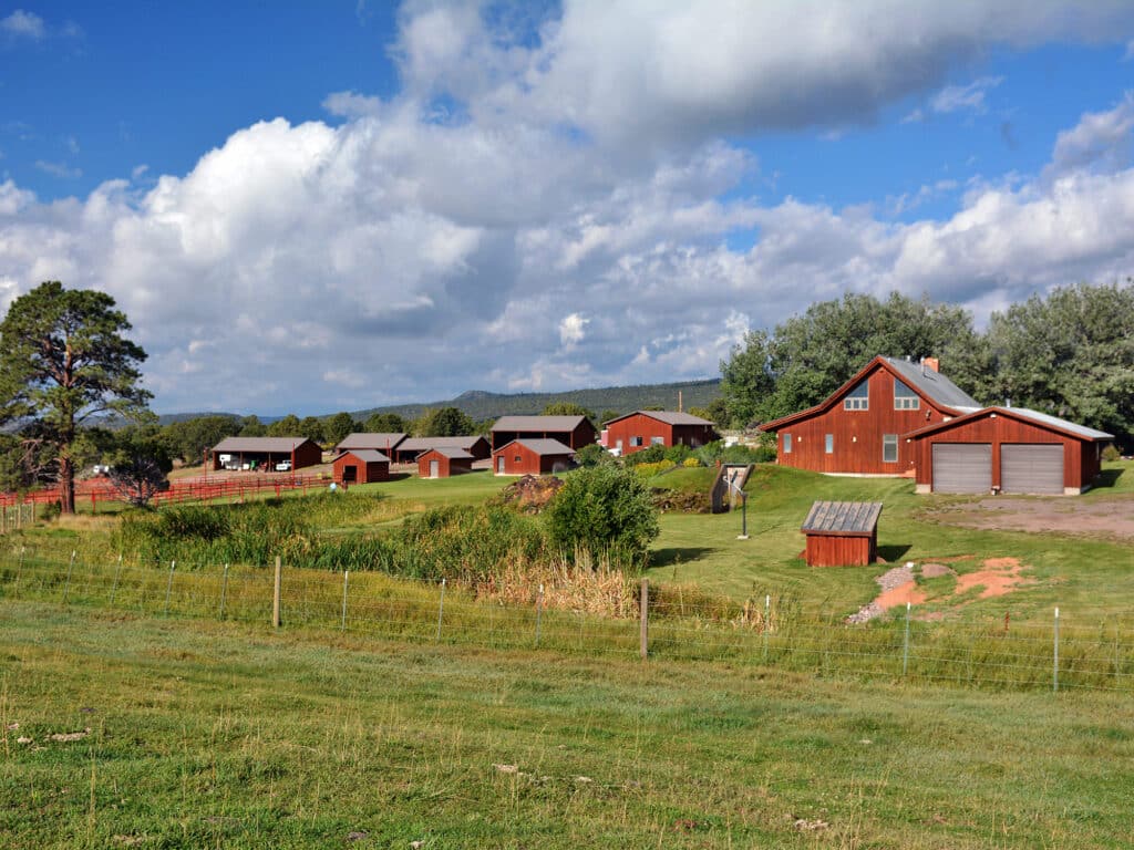 A cluster of red farm buildings, including barns and sheds, sits on green grass under a partly cloudy sky, surrounded by trees and a fence in a rural landscape—perfect as a ranch for sale or hunting property.
