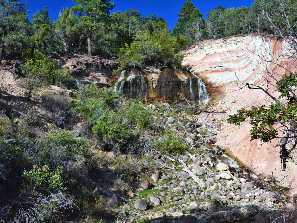 A small waterfall flows over layered rock formations, surrounded by green shrubs and trees under a clear blue sky—an inviting scene for those seeking land for sale or a picturesque cattle ranch. A rocky stream winds through the lush vegetation.