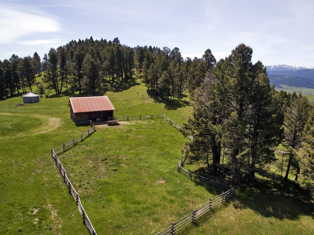Aerial view of a rustic wooden barn with a rusted roof, fenced pasture, and surrounding trees on a green hillside—perfect for a cattle ranch or recreational land—with mountains visible in the distance under a clear sky.