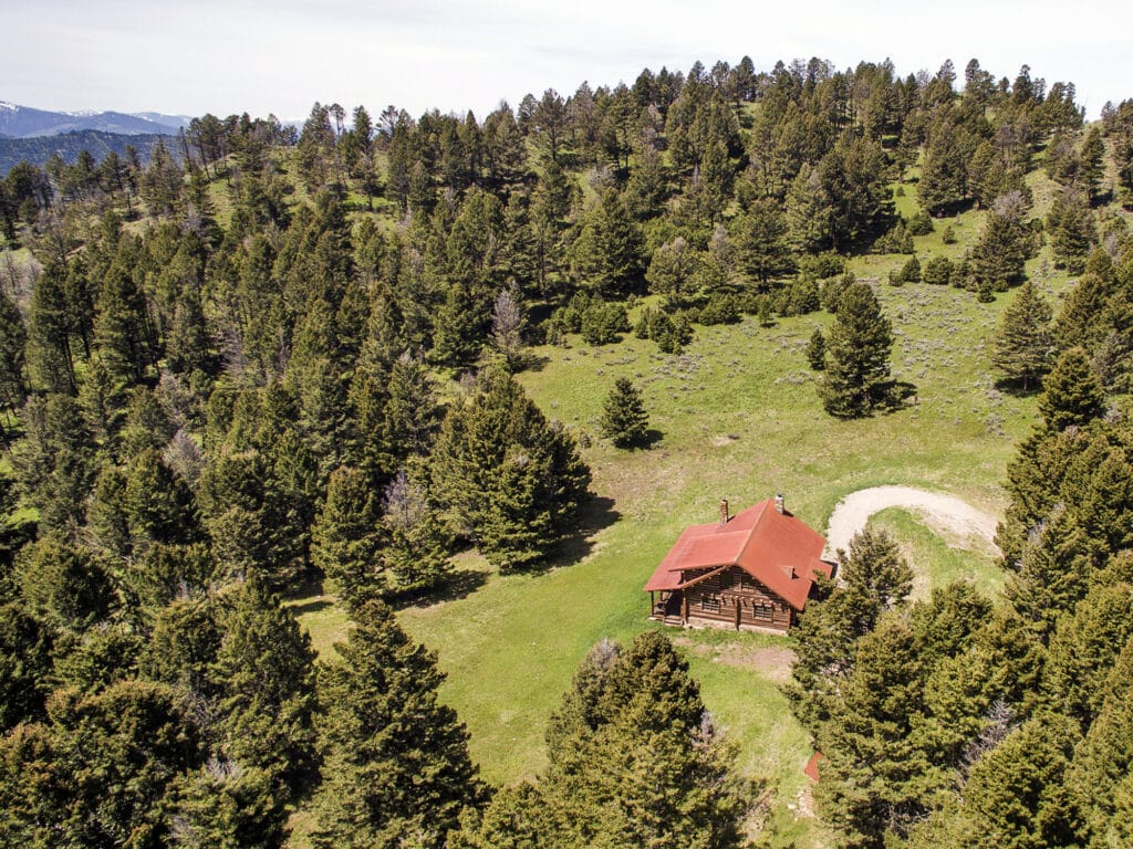 A small cabin with a red roof sits in a clearing surrounded by dense pine trees and green hills, viewed from above in a remote, mountainous area—perfect as hunting property or land for sale.
