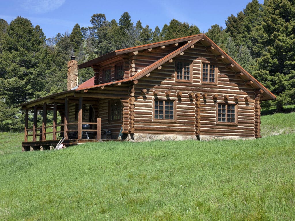 A rustic log cabin with a covered porch sits on a grassy hill, surrounded by dense green pine trees under a sunny blue sky—perfect as a hunting property or cozy ranch for sale.