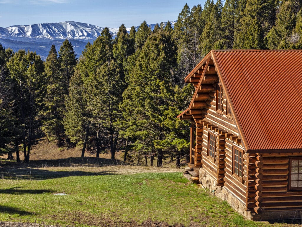 A rustic log cabin with a red metal roof sits on a grassy clearing surrounded by dense pine trees, perfect for recreational land or as part of a cattle ranch, with snow-capped mountains visible in the background under a clear blue sky.