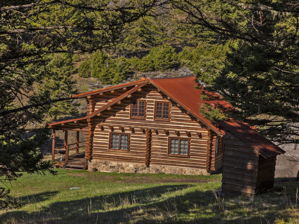 A rustic log cabin with a red roof sits on a grassy clearing surrounded by trees and hills, bathed in sunlight—an inviting hunting property perfect for outdoor enthusiasts.