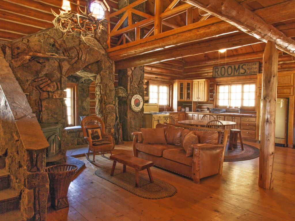 Rustic cabin interior with wooden beams, stone fireplace, leather sofa, wicker chairs, and kitchen area. Warm sunlight fills this inviting hunting property. A vintage Rooms sign hangs above the windows and kitchen cabinets.