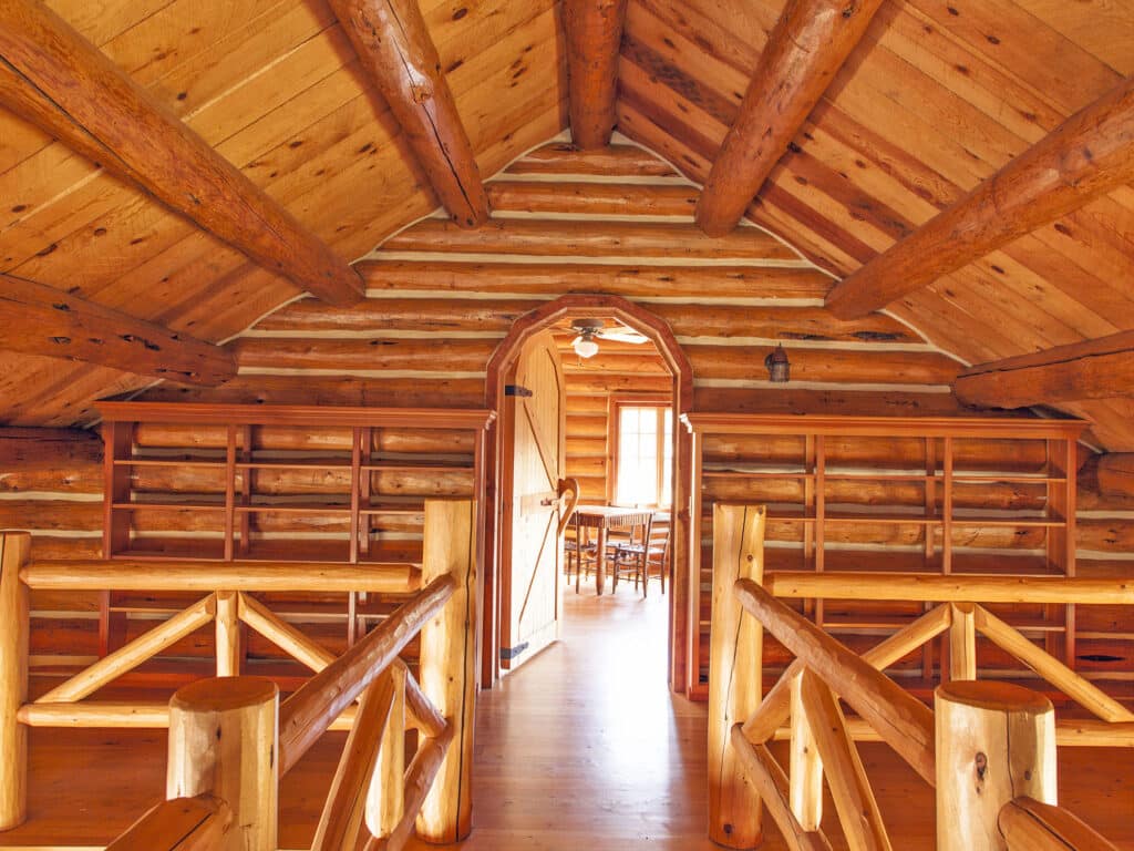 Interior of a rustic log cabin with wooden beams, shelves, and a sloped ceiling. An open doorway leads to a sunlit room with a table and chairs—perfect for those seeking unique ranch for sale or prime hunting property.