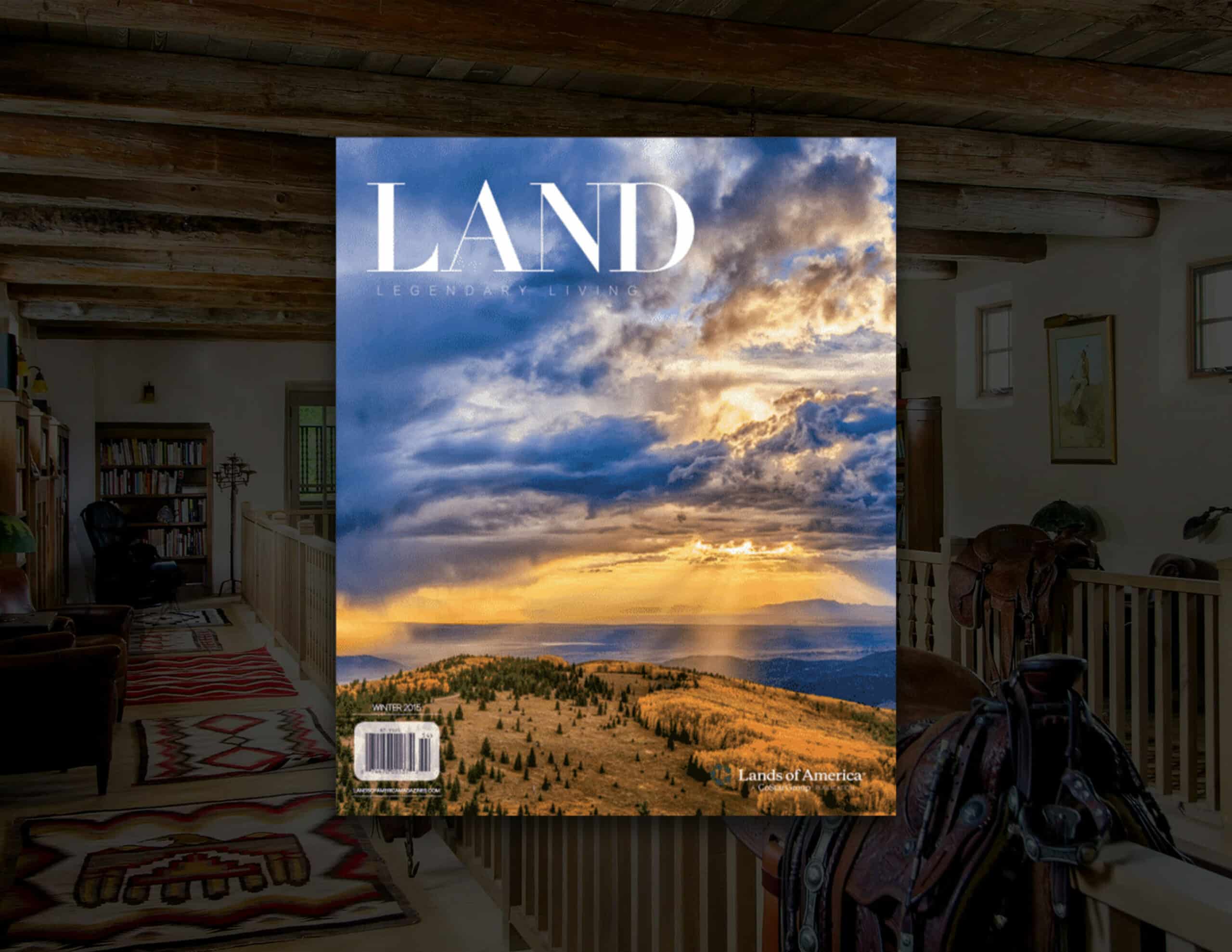 A magazine titled LAND features a dramatic sky over sunlit, hilly hunting property. It rests in the foreground of a rustic, wood-beamed room with western decor and a saddle in view.