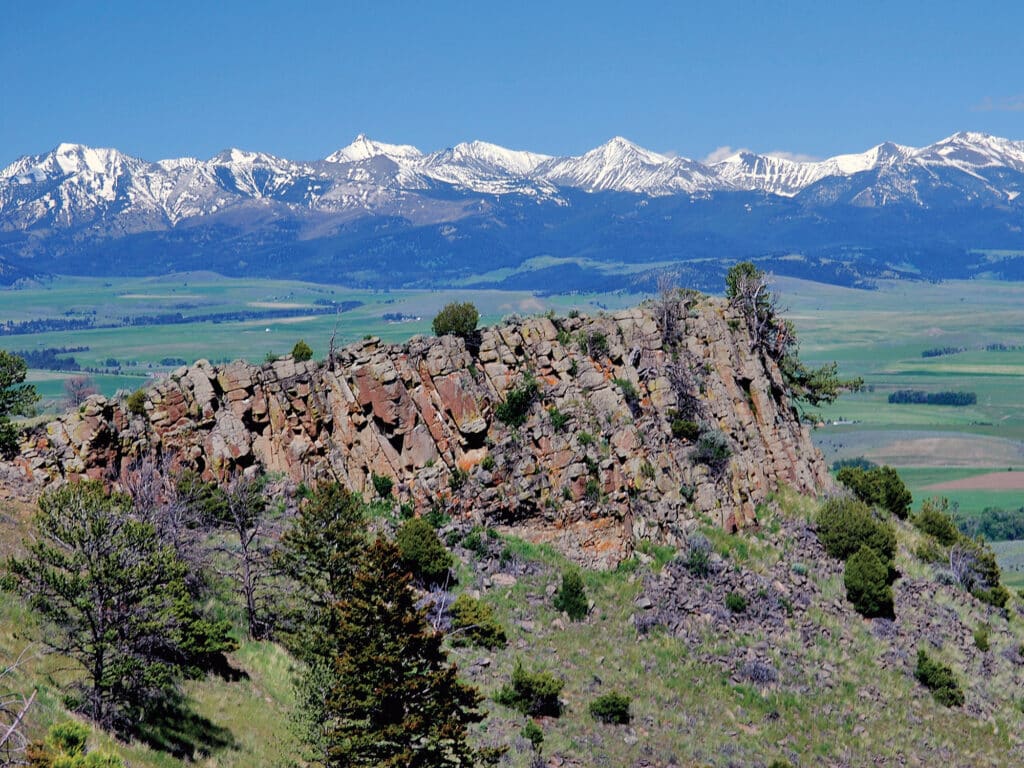 Rocky outcrop with sparse trees and shrubs in the foreground; green fields and a range of snow-capped mountains under a clear blue sky in the background, perfect for recreational land or a ranch for sale.
