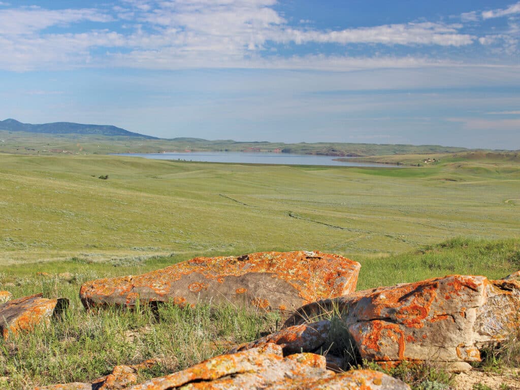 Large, orange-lichen-covered rocks sit in the foreground of a wide, grassy prairie landscape—an ideal setting for a cattle ranch. A calm lake stretches across the middle distance, with low hills and a blue sky with scattered clouds in the background.