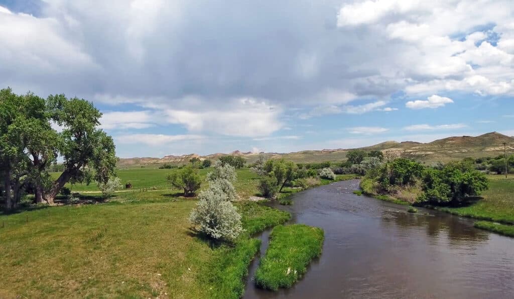 A winding river flows through a grassy landscape with scattered trees and rolling hills under a partly cloudy sky, making this ideal recreational land or hunting property.