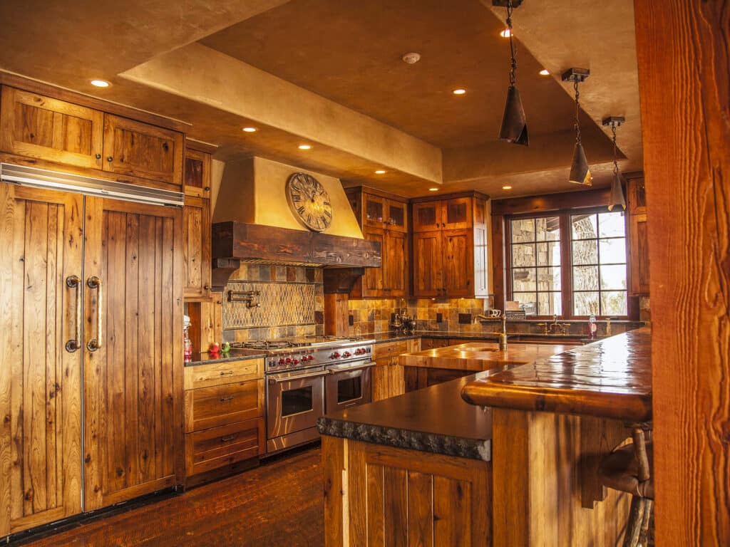 Warm, rustic kitchen with natural wood cabinets, a large wooden refrigerator, stone countertops, and a central island. Perfect for a cattle ranch home, the space features a range with decorative hood, large clock, and pendant lighting.