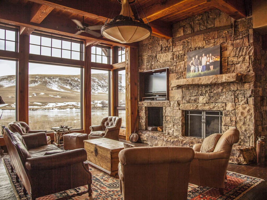 A cozy living room on a hunting property features leather chairs, a wooden coffee table, large windows framing a snowy landscape, and a stone fireplace with a photo above it. Warm light and rustic decor create a welcoming atmosphere.