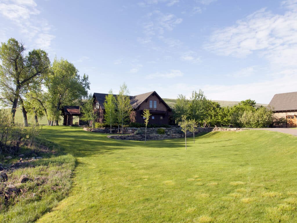 A rustic wooden house sits among trees and shrubs on a large green lawn under a blue sky with scattered clouds. Ideal as recreational land or hunting property, another building and a small wooden structure are visible in the background.